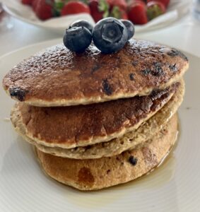 A stack of three golden-brown Lemon Blueberry Pancakes topped with fresh blueberries, drizzled with maple syrup, and served on a white plate, with strawberries blurred in the background.